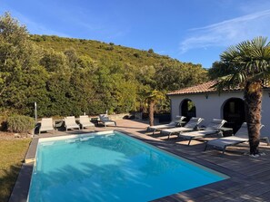Una piscina con sillas de descanso, un pequeño edificio con arcos y una colina al fondo.