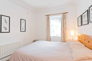 A hotel room with a large bed, a radiator, a window with curtains, and framed artwork on the walls.