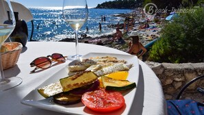 A table with a plate of grilled vegetables and a glass of white wine, overlooking a beach with people and a clear blue sea. - "BUENA VISTA" 55m2, south-facing balcony, fenced, 5ft. beach, suitable for children, Rovinj (Rovinj)