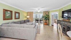 A living room with a grey sectional sofa, a dining area with a table and chairs, a TV on a cabinet, and a large potted plant. - Heron 408 — 3BR Bayside Condo at Destin West with Heated Pool (Fort Walton Beach)