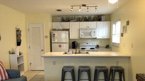 Cocina compacta con gabinetes blancos, microondas integrado, estufa y refrigerador. Hay cuatro taburetes en la isla de la cocina y una mesa de centro en la sala.