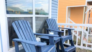 Dos sillas Adirondack azules sobre un muelle de madera con baranda blanca, frente a un edificio con revestimiento naranja.