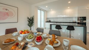 Un moderno comedor con una mesa de madera preparada para el desayuno, incluyendo cruasanes, frutas y bebidas.