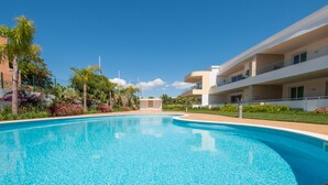 Una piscina con superficie de agua cristalina y azul, rodeada de un jardín bien cuidado con plantas variadas y un edificio moderno con balcones al fondo.