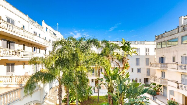 A courtyard with a fountain, palm trees, and a white building with balconies. - Appartamento Corso Italia II by Barbarhouse (Gallipoli)