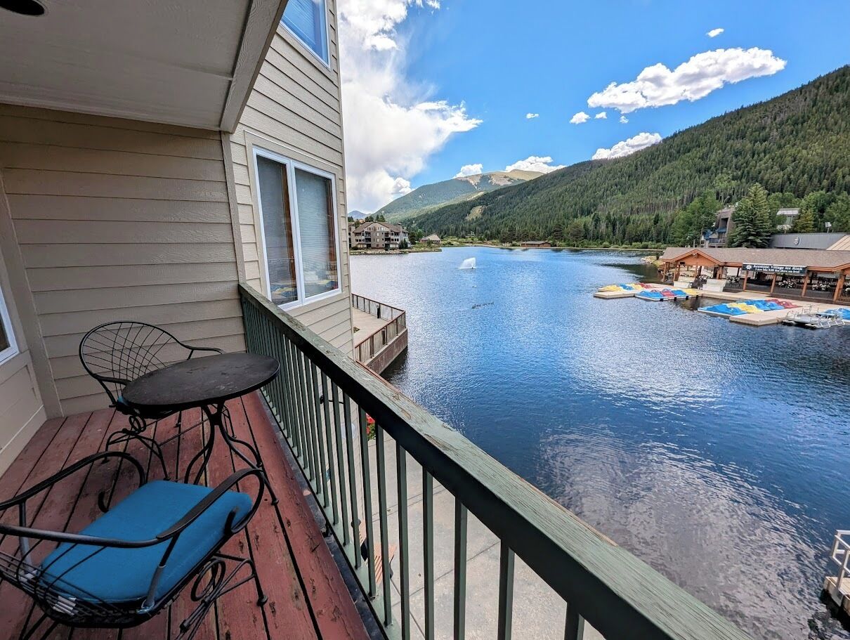 A balcony with a table and chairs overlooking a lake, with buildings and mountains in the background.