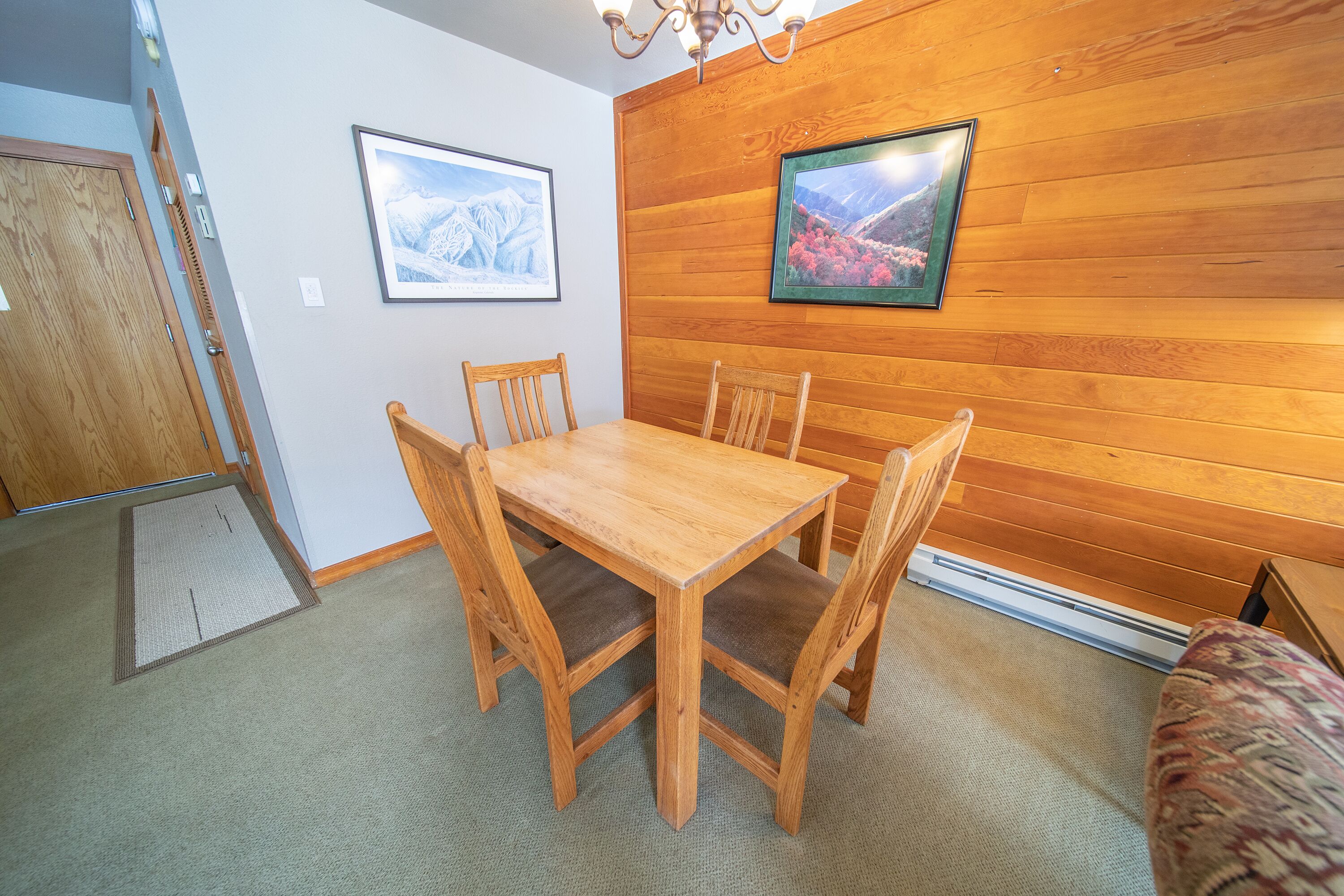 A wooden-paneled room with a dining table and chairs, two framed pictures on the wall, and a carpeted floor.
