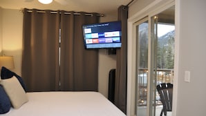 Hotelkamer met een bed, een aan de muur bevestigde televisie, een raam met uitzicht op bomen en bergen, en een stoel buiten.