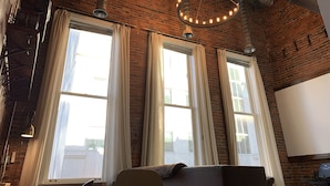 Een kamer met zichtbare bakstenen muren, grote ramen met witte gordijnen, een ronde lamp en een moderne bank.