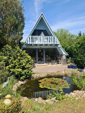 Un bâtiment de forme triangulaire doté d’un balcon, entouré d’un étang orné de nénuphars et de rochers.