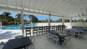 A covered outdoor seating area with tables and chairs, a pool, and palm trees in the background. - Coconut Castaway | Ocean Views, Golf Cart & Club Pool (Upper Captiva)