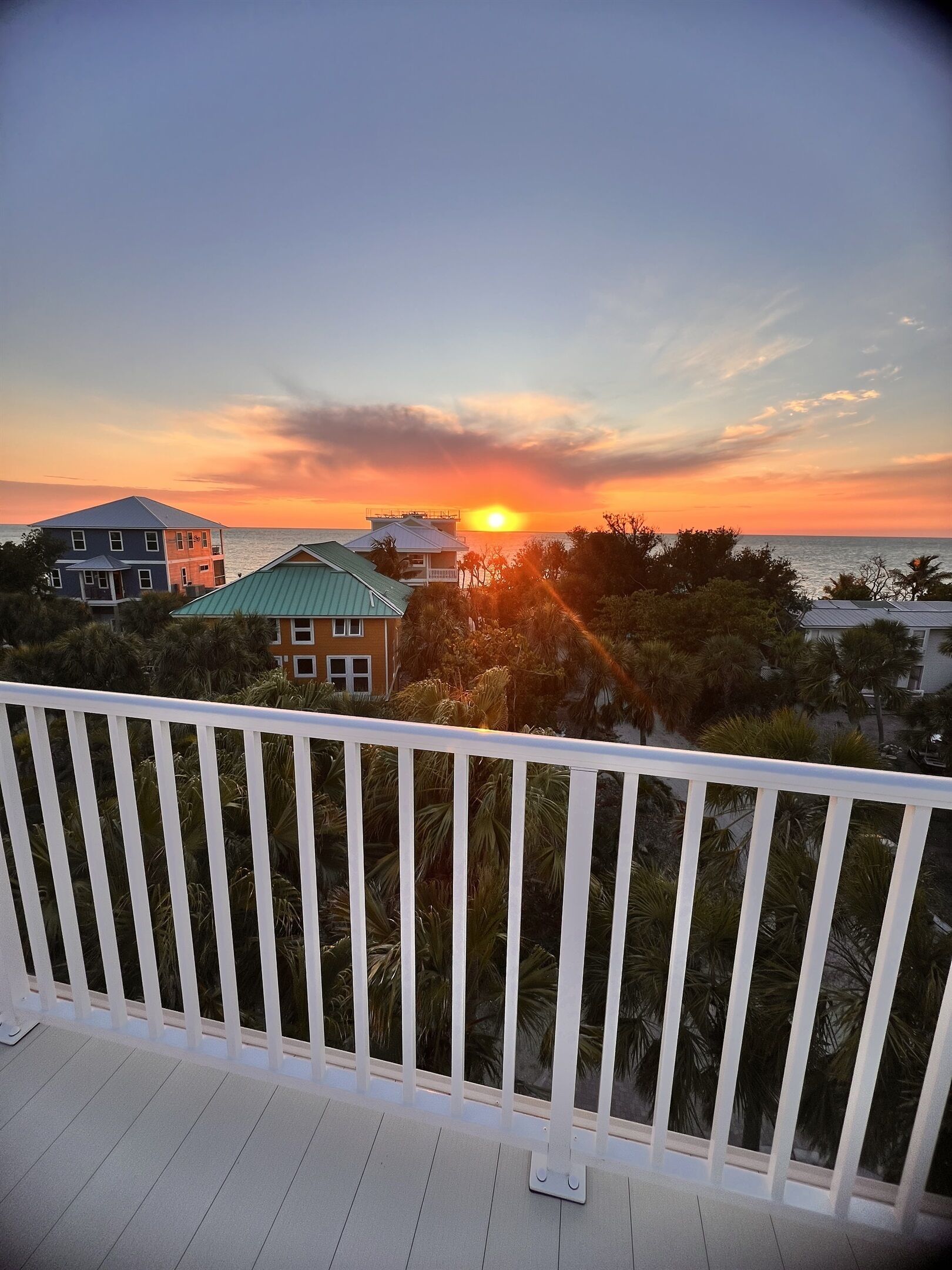 A balcony with a view of a sunset over the ocean, palm trees, and buildings in the background.
