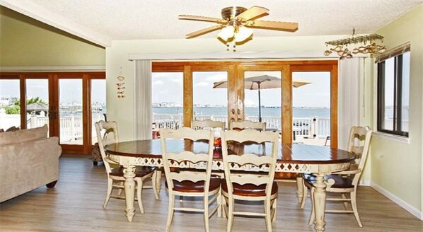 A dining area with a round table and chairs, large windows with a view of the sea, and a ceiling fan.