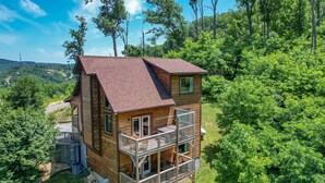 A wooden cabin with a balcony, surrounded by lush greenery and hills.