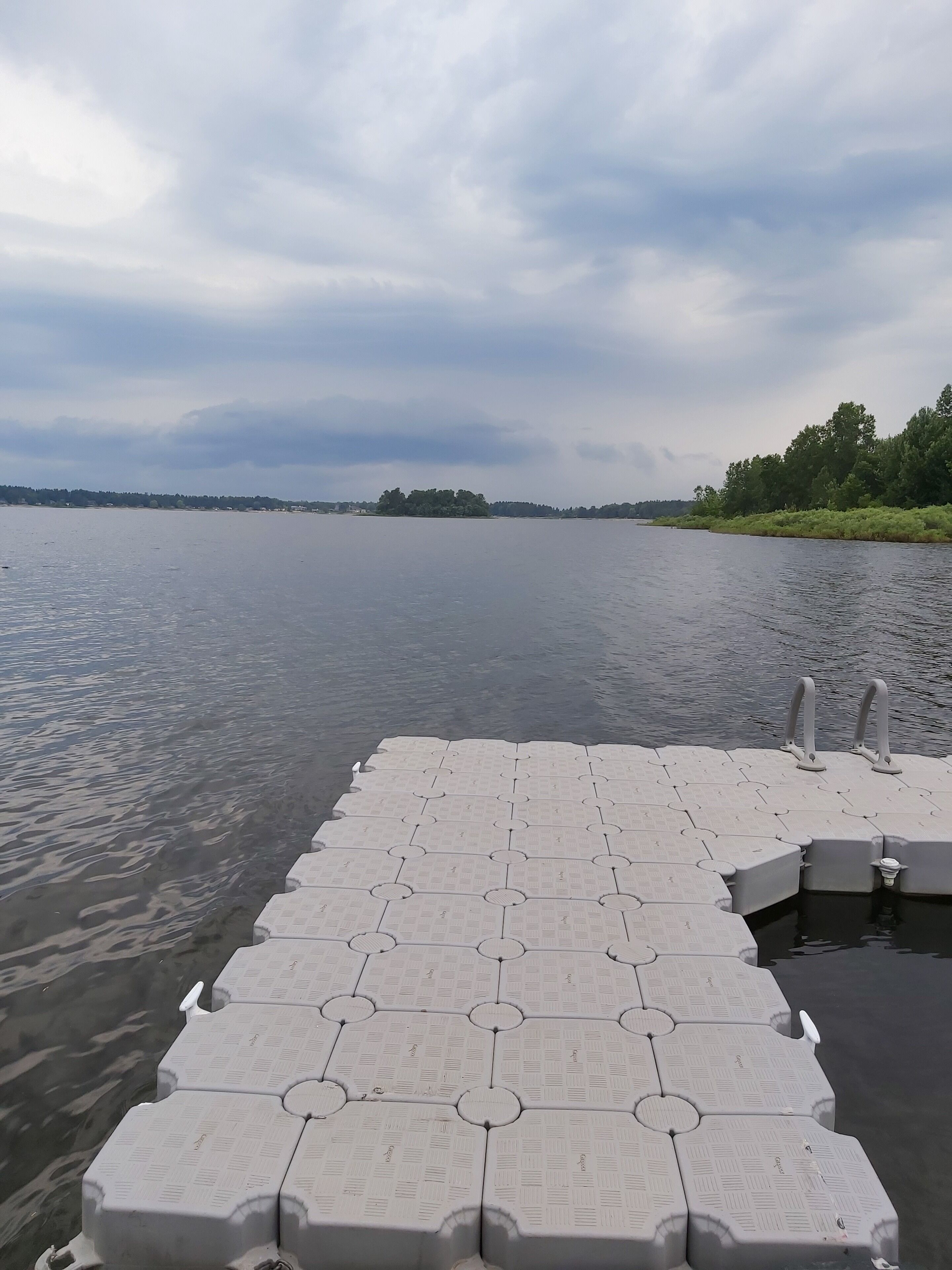 Eine Betonsteg mit Gittermuster und Metallgeländern, der sich über ruhiges Wasser zu einer fernen Uferlinie und Bäumen erstreckt.