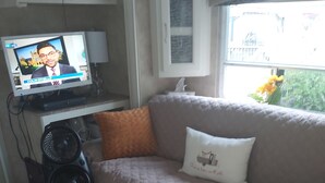 A living room with a beige sofa, a white ottoman, a television displaying a news program, and a window with a view of greenery. - Little Happy Place (Belwood)