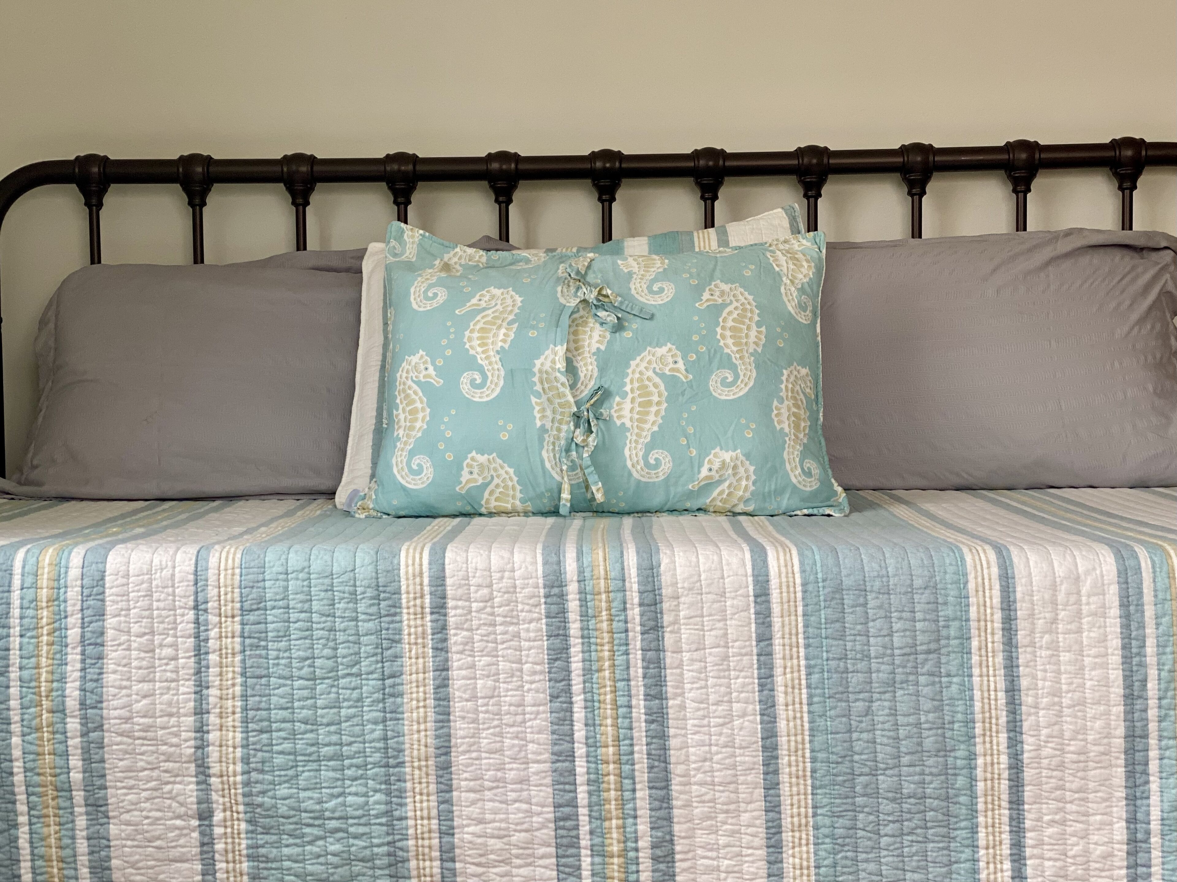 A bed with a striped quilt, a teal pillow with seahorse patterns, and two grey pillows.