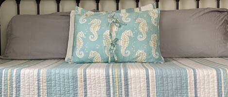 A bed with a striped quilt, a teal pillow with seahorse patterns, and two grey pillows.