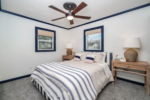 Quarto com cama, duas mesas de cabeceira, ventilador de teto e duas janelas com persianas.