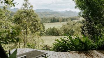 Une terrasse en bois avec une chaise, offrant une vue sur un paysage luxuriant, avec des arbres et des montagnes à l’horizon.