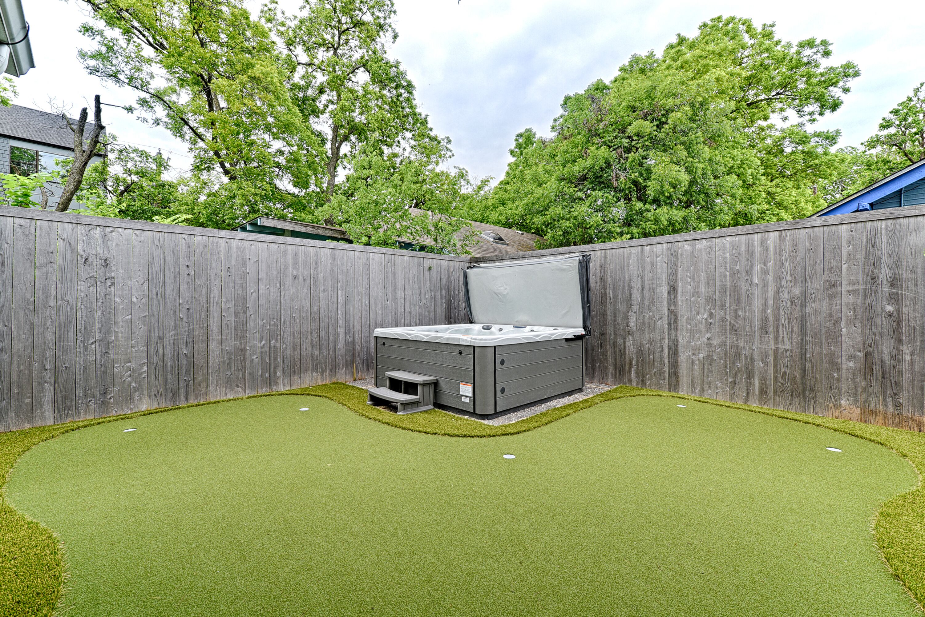 A hot tub in a backyard with artificial grass, surrounded by a wooden fence and trees.