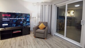 Een woonkamer met een grote flatscreen-tv waarop de Netflix-interface te zien is, een grijze fauteuil met een geel kussen, een zwarte entertainmentkast met een spelcomputer, en een schuifdeur die leidt naar een balkon met lichtslingers.