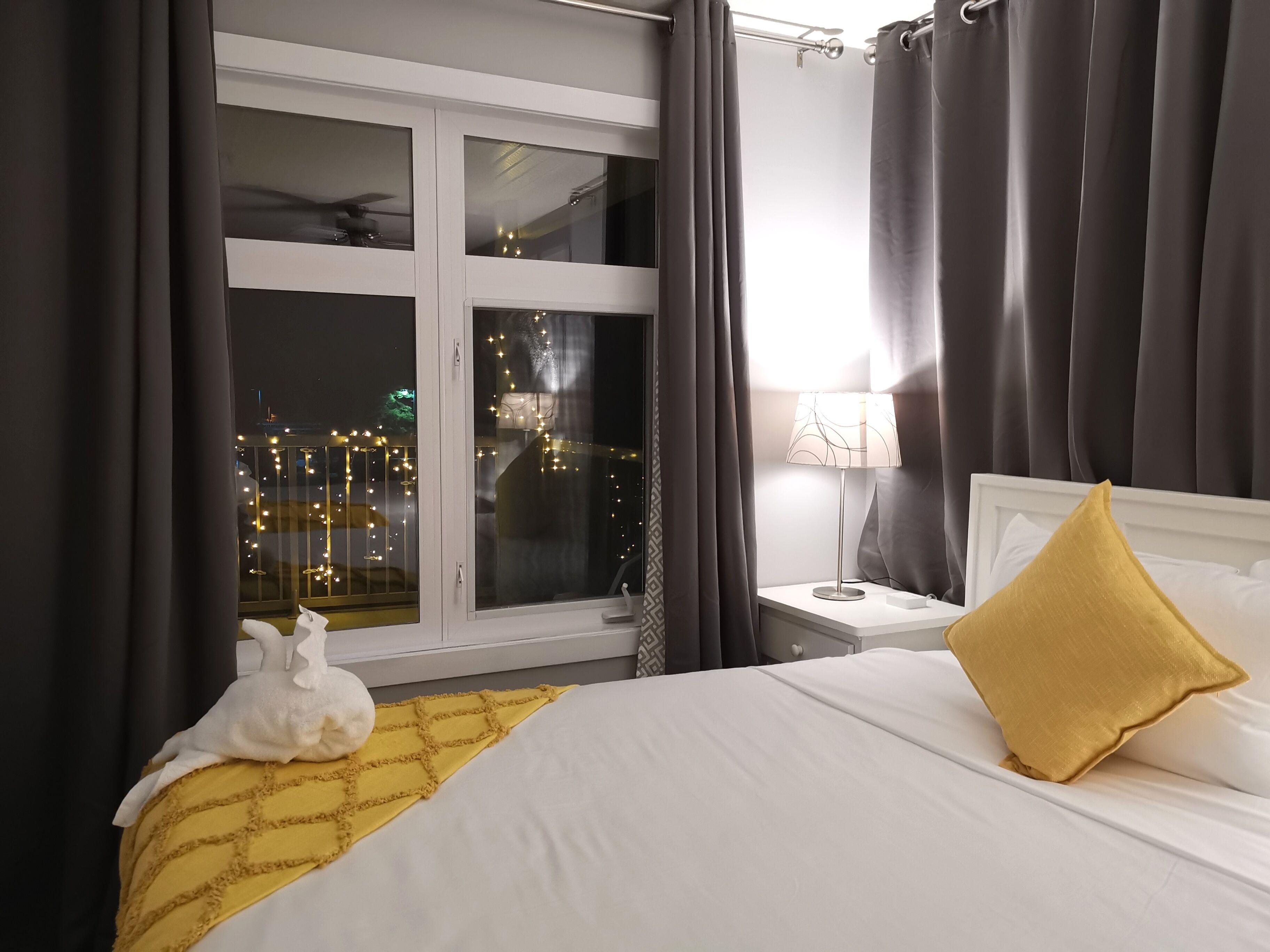 A hotel room with a bed, a nightstand, a lamp, a window with curtains, and a view of the city lights outside.