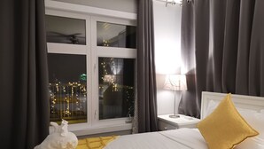 Een hotelkamer met een bed, nachtkastje, lamp, raam met gordijnen en uitzicht op de lichtjes van de stad.