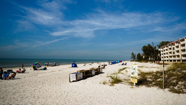 A beach with tents and beach chairs, a wooden fence, and a building with multiple floors. - Beautiful Townhome Steps from Beach (Indian Shores)