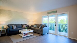 A living room with a grey sofa, a white coffee table, and a bookshelf. There is a sign that reads "Beach House" on the wall. - Beautiful Townhome Steps from Beach (Indian Shores)