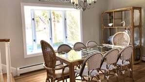 Comedor con mesa de vidrio, sillas de mimbre, una araña y un ventanal con vistas a árboles.