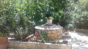 A stone fountain surrounded by lush greenery and flowering plants in a garden setting. - Yria suite (Katastari, Allykes)