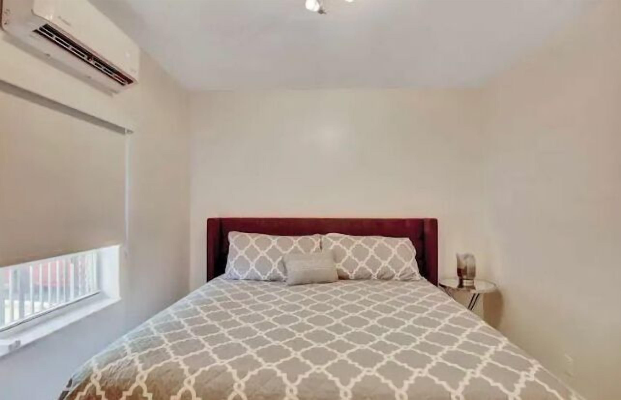 Quarto com cama, mesa de cabeceira, luminária, janela com persianas e ar condicionado.
