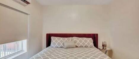 Quarto com cama, mesa de cabeceira, luminária, janela com persianas e ar condicionado.