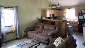 Una sala de estar con un sofá marrón, una mesa de centro con revistas y un televisor. Se ve una cocina con armarios de madera y un refrigerador.
