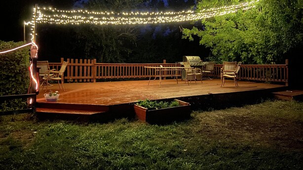 Una terraza al aire libre con barandilla de madera, luces colgantes y mobiliario exterior.