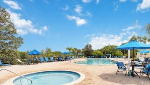 Ein Poolbereich mit Liegestühlen und Sonnenschirmen, umgeben von Bäumen und blauem Himmel.