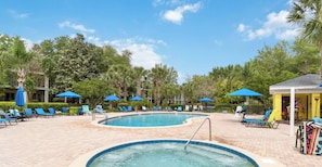 Un espace piscine doté de chaises longues et de parasols, entouré d’arbres et d’un bâtiment.