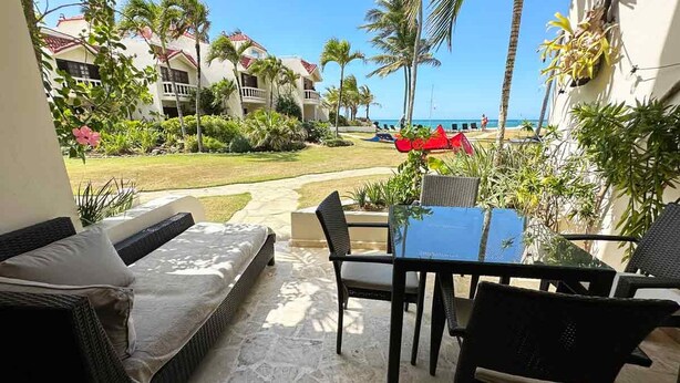 A patio with a sofa, table, and chairs, overlooking a pool and a beach with palm trees and a building in the background.