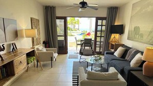 A modern living room with a sofa, armchairs, a coffee table, and a TV. There is a balcony with a view of the outdoors.