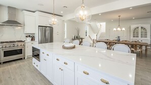 Een moderne keuken met witte kasten, een groot eiland, roestvrijstalen apparatuur en hanglampen.