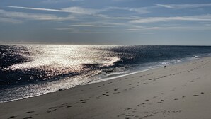 Een strand met voetsporen die naar het water leiden, een heldere lucht en zonlicht dat op de oceaan reflecteert.