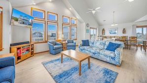 A spacious living room with a large window, a ceiling fan, and a dining area with wooden furniture. - Semi-Oceanfront Dog Friendly w/ Pool, Hot Tub, Game Room, Theater, Tiki Bar (Waves)