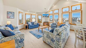 A spacious living room with a high ceiling, large windows offering views of the surrounding area, and a dining area with a table and chairs. - Semi-Oceanfront Dog Friendly w/ Pool, Hot Tub, Game Room, Theater, Tiki Bar (Waves)