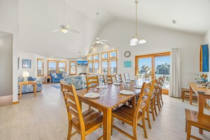 Un vaste espace de vie comprenant une table à manger pour six personnes, entourée de chaises en bois. On y trouve deux ventilateurs de plafond, une grande fenêtre donnant sur des maisons et une horloge au mur.