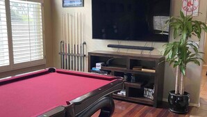 A pool table with a red felt surface, a television mounted on the wall, and a potted plant to the right. - 🔥HEATED Pool, Pool Table, Golf, and Spring Training (Glendale)