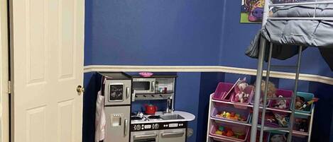 A room with a bunk bed, a kitchenette with a microwave and stove, and a toy shelf.