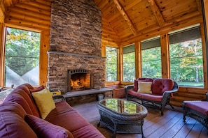 A cozy cabin interior with a stone fireplace, wicker furniture, and large windows offering a view of the forest. - New Large Adirondack Lodge with Pond Waterfront and A/C (Old Forge)