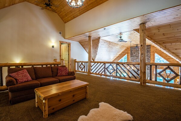 Un vaste espace de vie de style loft, doté d’un plafond haut, de poutres apparentes, d’un canapé en bois avec des coussins rouges, d’une table basse en bois et offrant une vue sur l’extérieur grâce à un balcon agrémenté d’une balustrade en bois.