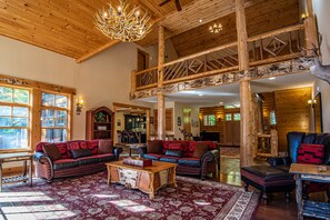 A spacious lodge with a high ceiling, wooden beams, and a grand chandelier. There are two sofas with red and black cushions, a wooden coffee table, and a large rug. - New Large Adirondack Lodge with Pond Waterfront and A/C (Old Forge)